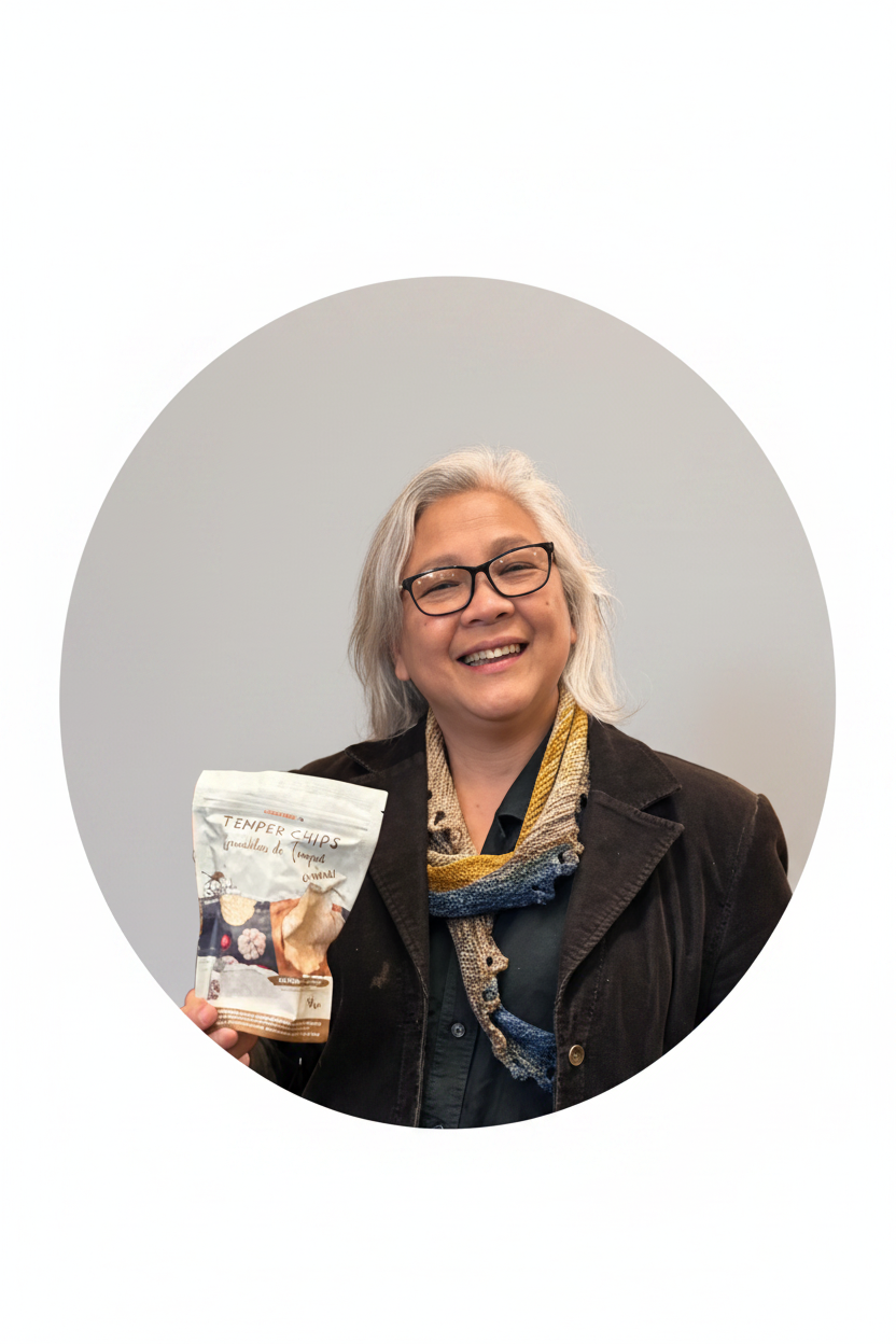 A smiling woman with gray hair and glasses stands in a grocery store aisle holding a bag of Envy Farms Organic Sweet Potatoes Chips. Shelves behind her are stocked with various snack bags.